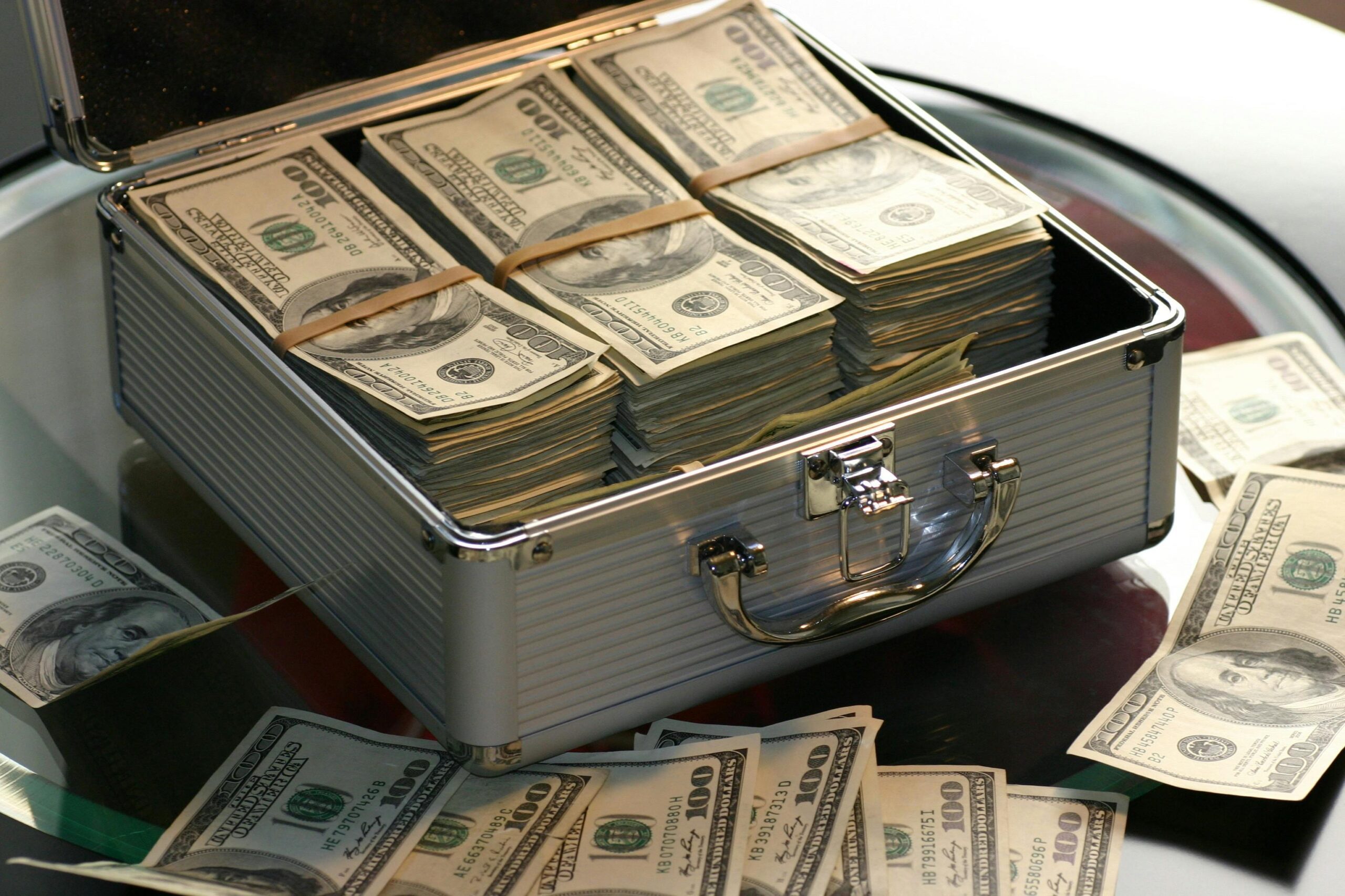 Best Apps to Manage Money Open briefcase filled with stacks of hundred dollar bills on a glass table, representing wealth.